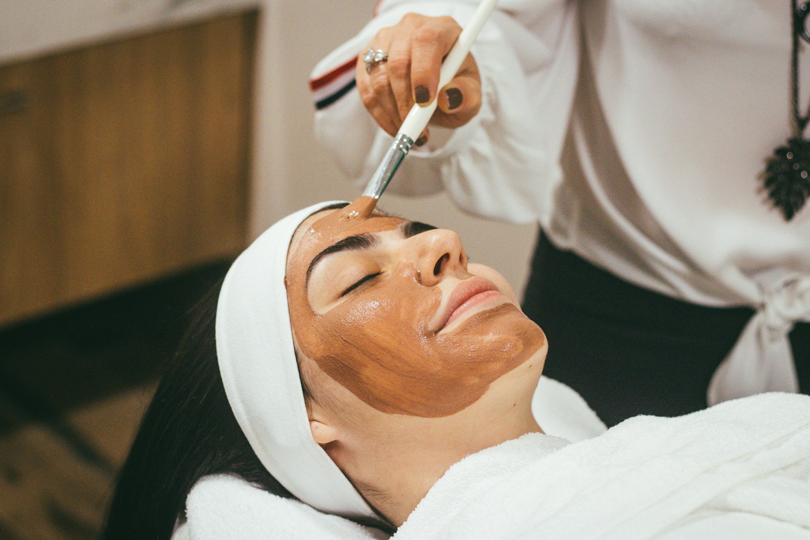 An aesthetician brushing a brown clay mask onto a client's face at a treatment table