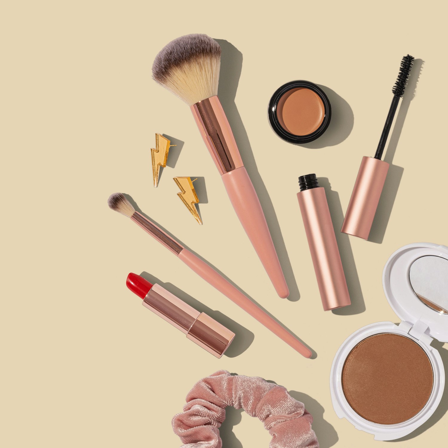 Pink and rose makeup items arranged on a pale sand background, including brushes, lipstick and a compact