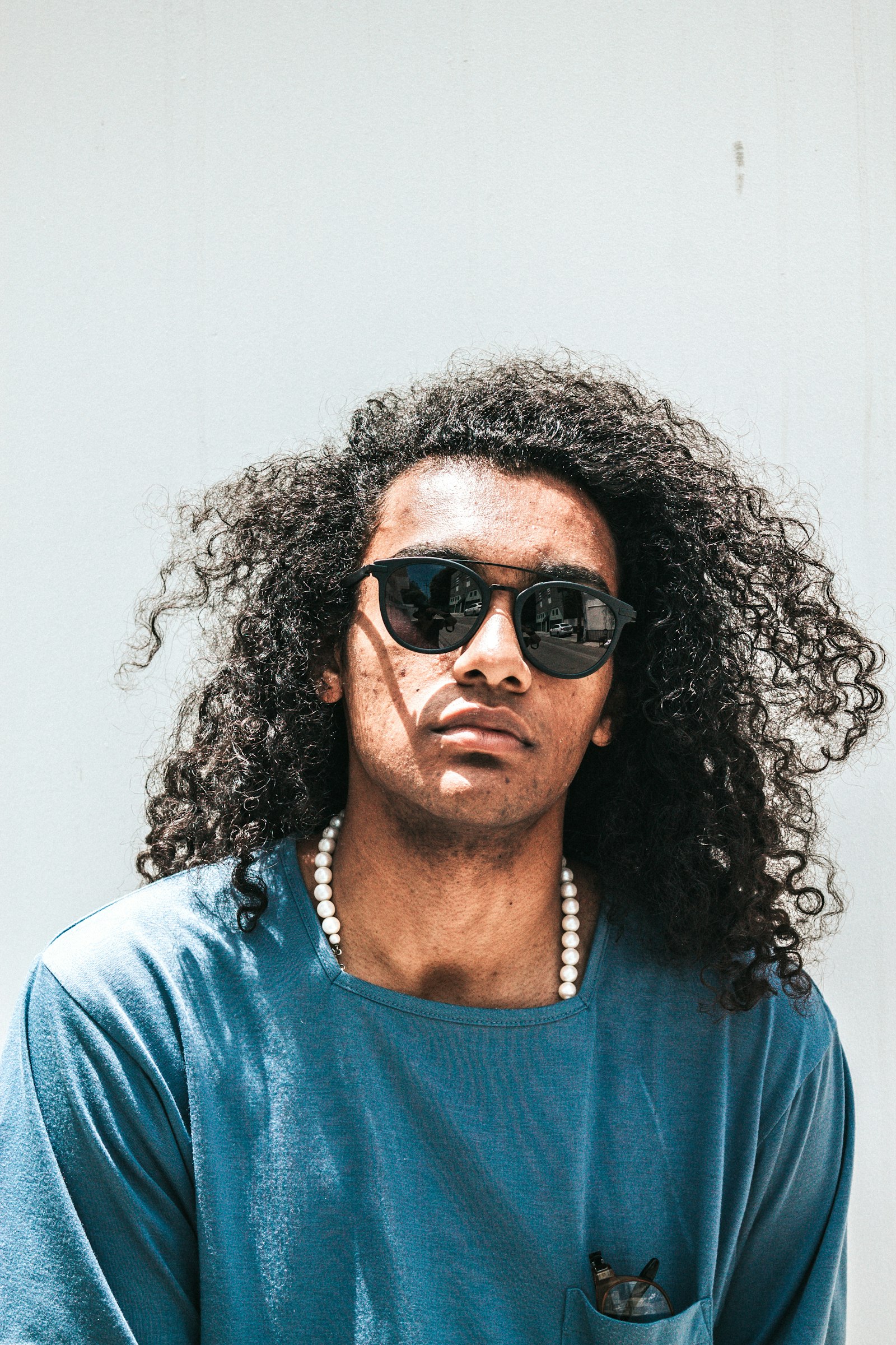 A portrait of a person with long curly dark hair wearing sunglasses against a pale wall