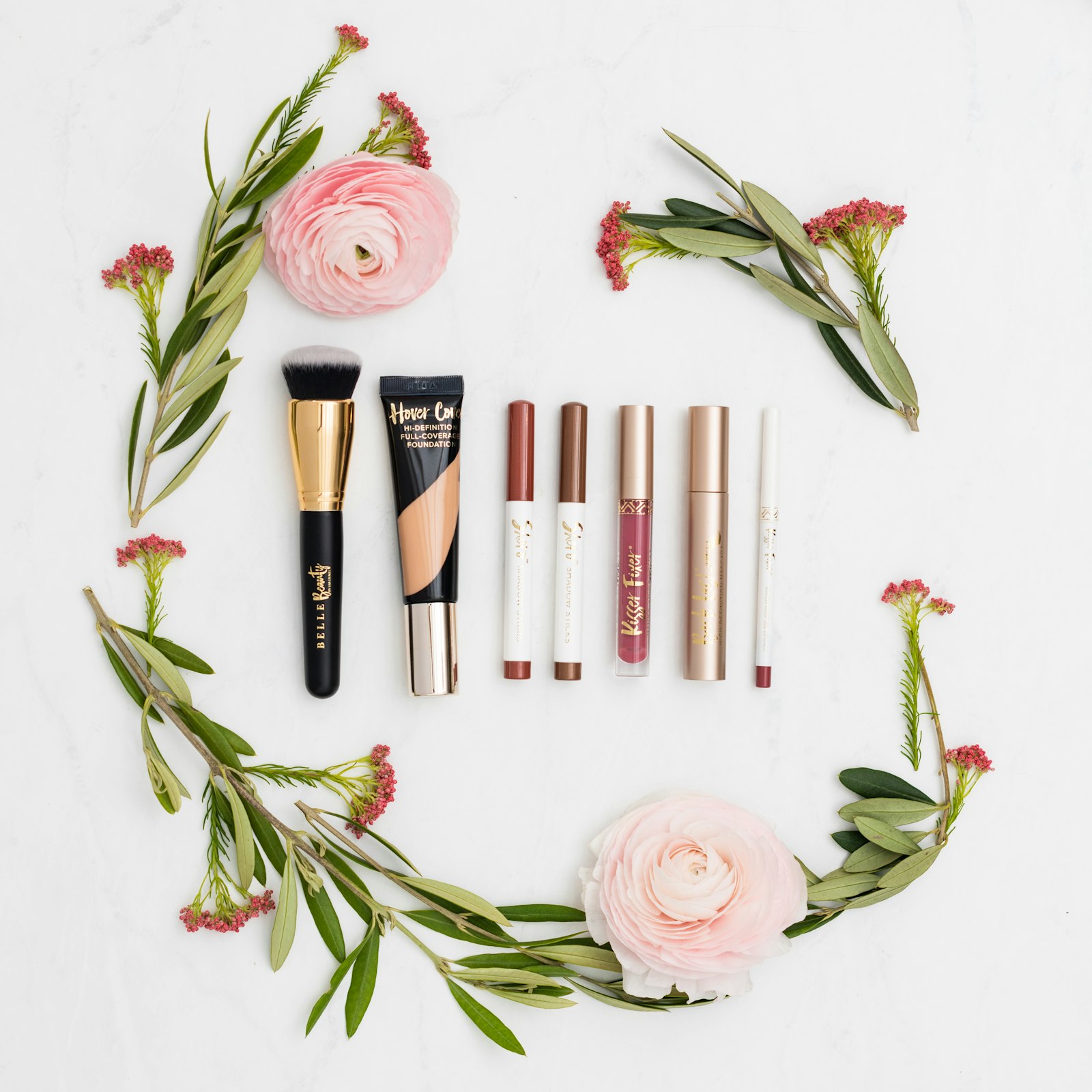 Cosmetics arranged in a circle with pink ranunculus flowers on a white surface